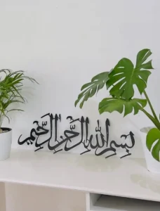 The best Arabic calligraphy art in black metal featuring the phrase ‘بسم الله الرحمن الرحيم’ (In the name of Allah, the Most Gracious, the Most Merciful), displayed beautifully on a white shelf between two vibrant green indoor plants. A perfect Islamic decor piece for a peaceful and modern home setting. best arabic calligraphy art