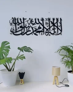 Best Arabic calligraphy art in black metal featuring the Shahada ‘لا إله إلا الله محمد رسول الله,’ beautifully mounted on a clean white wall. Surrounded by elegant green plants, a minimalist wooden table lamp, and a decorative incense burner—perfect for Islamic home decor and modern interior design.