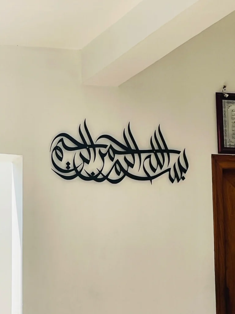 The best Arabic calligraphy art in elegant black metal featuring the phrase ‘بسم الله الرحمن الرحيم’ (In the name of Allah, the Most Gracious, the Most Merciful), beautifully mounted on a light cream wall near a wooden door. A stunning piece of Islamic wall decor for a modern and peaceful home interior. best arabic calligraphy art